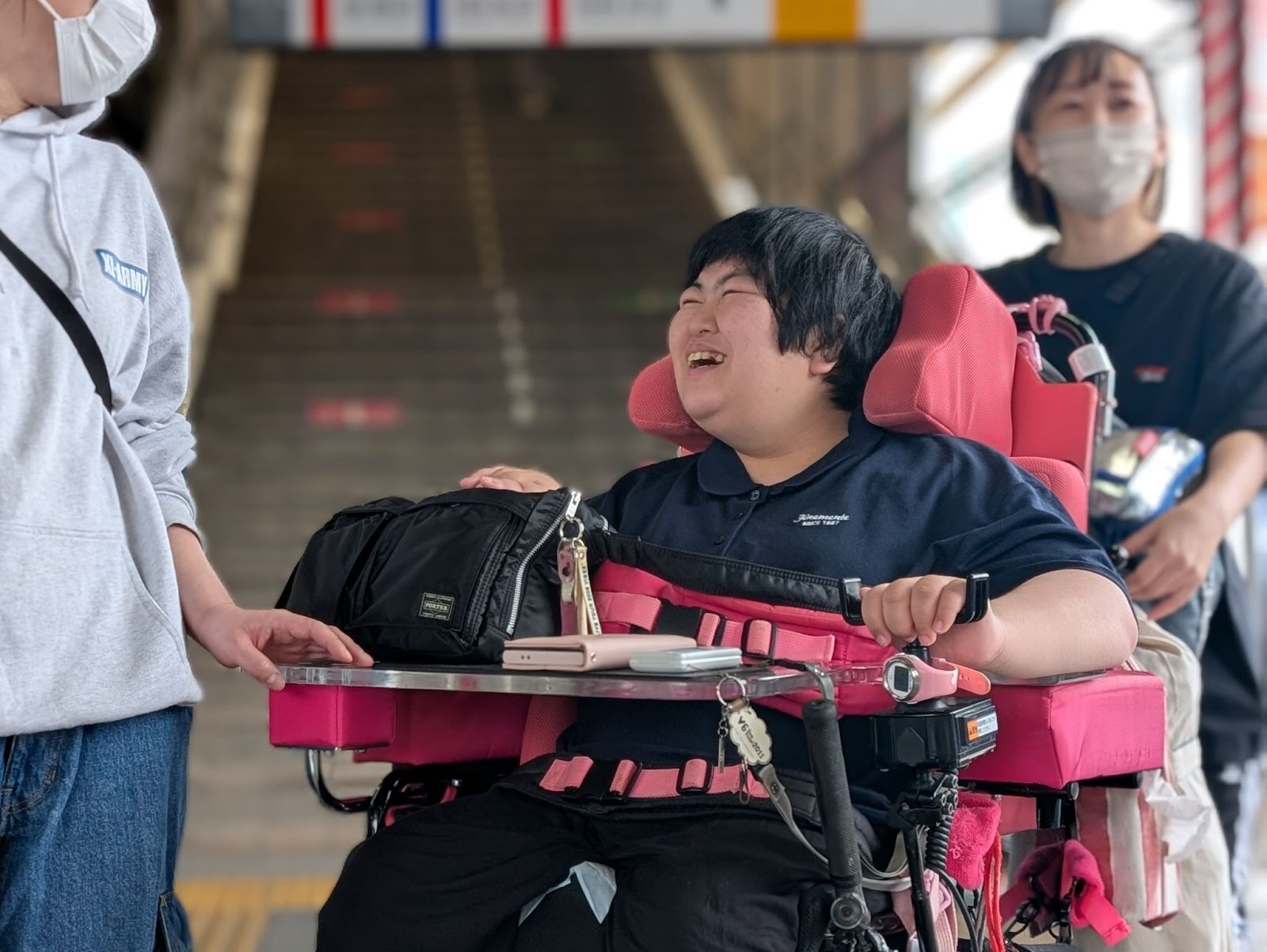 笑顔で過ごす重度障害者の若者が車椅子に座り、側にはサポートしているスタッフがいる様子。背景には階段と駅の構造物が見える。