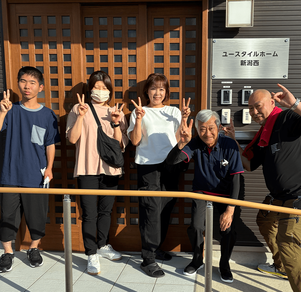 ユースタイルラボラトリーの新潟西のスタッフがポーズを取っている写真。5人が笑顔で並び、指でVサインをしています。背景には施設の入り口が見えます。
