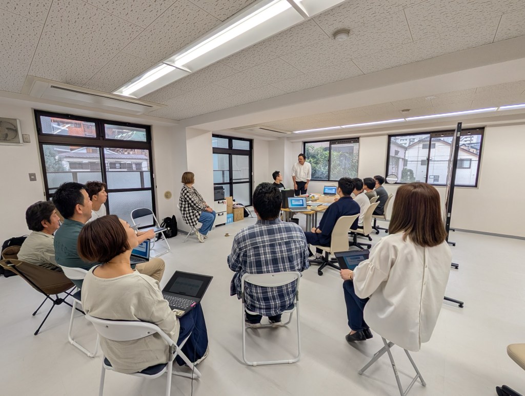 ビジネスミーティングを行う参加者たちが集まっている室内の様子。テーブルにはパソコンが置かれ、数人のメンバーが講師の話を聞いている。