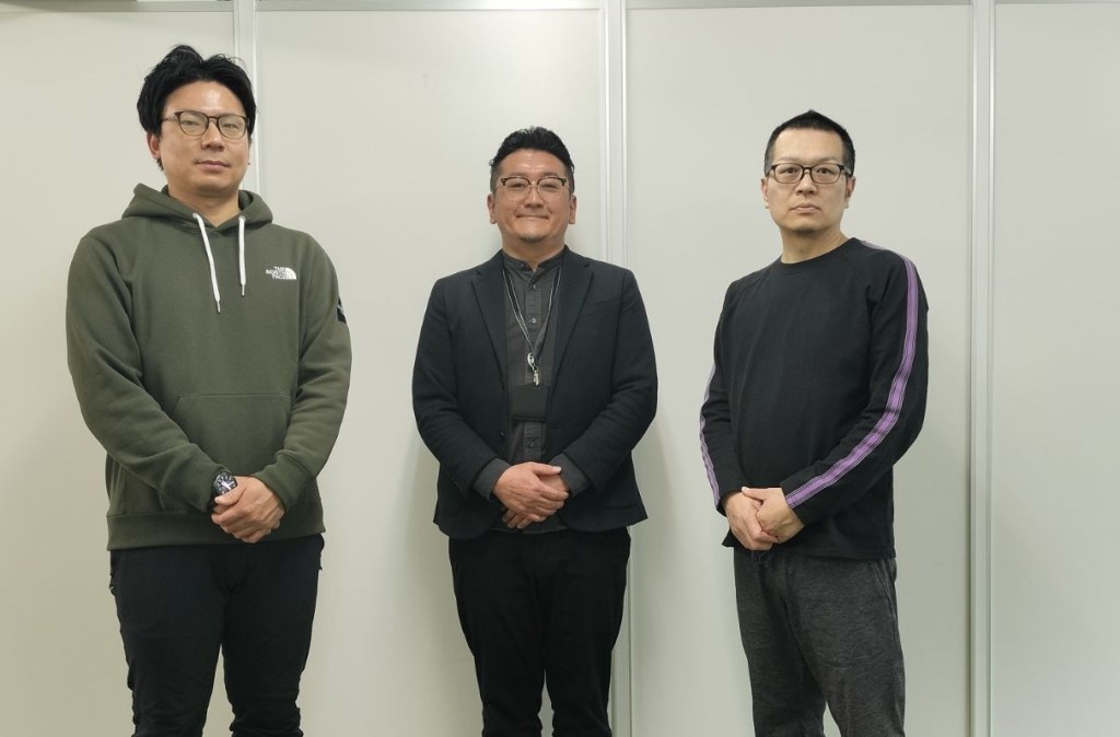 3人の男性が立っている写真。左から順に、緑のフード付きジャケットを着た男性、黒のスーツを着た男性、黒のTシャツを着た男性で、背後に白い壁がある。