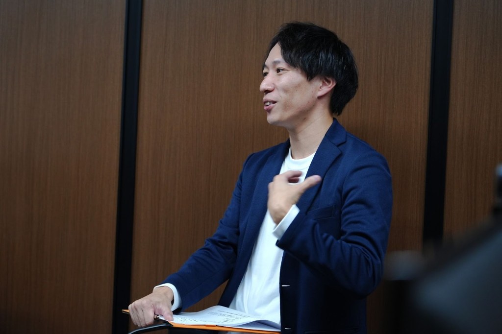 会社の会議室で内定式で話している男性の姿。背景は木目調の壁でアットホームな雰囲気が漂う。